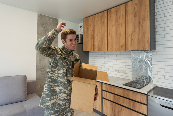 Uśmiechnięty mężczyzna w kamuflażu trzymający kartonowe pudełko w nowych apartamentach. - Zdjęcie, obraz