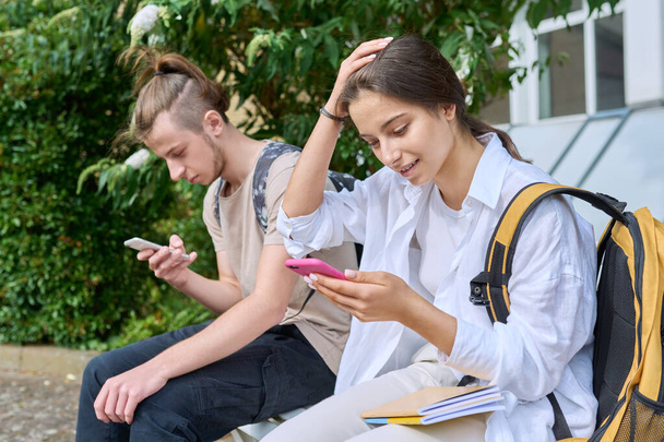 Teenage high school students, guy and girl, sitting on bench with backpacks, near academic building, using smartphones. Adolescence, youth, education, lifestyle, technology concept - Foto, afbeelding