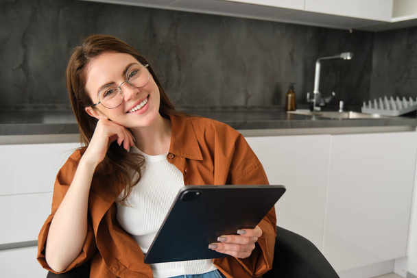 Portrait of beautiful smiling girl, wearing glasses, sitting in kitchen with digital tablet, working on remote from home. - Photo, Image