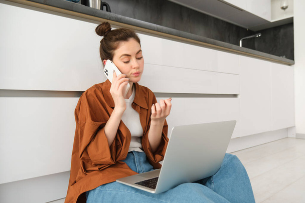 Mujer que parece aburrida y sin diversión mientras está sentada en el suelo y escuchando la conversación, tiene un ordenador portátil en vueltas, esperando en línea, haciendo una llamada telefónica. - Foto, Imagen