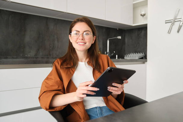 έννοια της ηλεκτρονικής μάθησης και του απομακρυσμένου χώρου εργασίας. Νεαρή γυναίκα με γυαλιά, φοιτήτρια που σπουδάζει στο σπίτι στην κουζίνα, διαβάζει σε ψηφιακό tablet, κοιτάζοντας το έργο της, συνδέεται με online συνάντηση. - Φωτογραφία, εικόνα