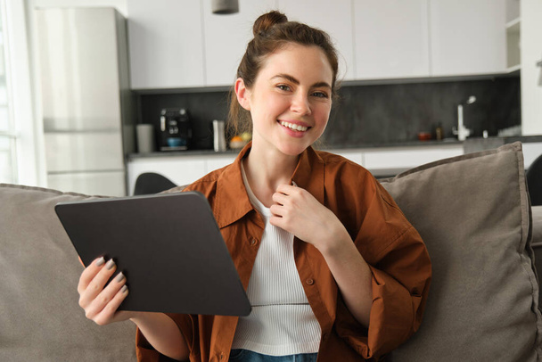 Schattige jonge brunette vrouw met digitale tablet in de hand, zitten op de bank, glimlachen en lachen op de camera. - Foto, afbeelding