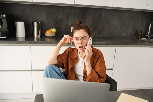 Πορτρέτο της γυναίκας με σοκαρισμένο πρόσωπο, λαμβάνει σχετικά με άσχημα νέα από την τηλεφωνική συνομιλία, κάθεται στο σπίτι με φορητό υπολογιστή, εργασίας, μιλώντας με κάποιον στο τηλέφωνο. - Φωτογραφία, εικόνα