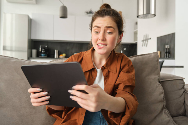 Portrét mladé ženy, která vypadá vystresovaně a napjatě na digitálním tabletu, hraje obtížnou videohru, sedí na gauči, používá gadget, tráví čas doma. - Fotografie, Obrázek