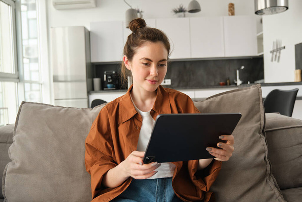 Porträt einer jungen Frau, die auf der Couch im Wohnzimmer sitzt, Filme auf dem digitalen Tablet anschaut, auf dem Gerät liest, das Wochenende zu Hause genießt. - Foto, Bild
