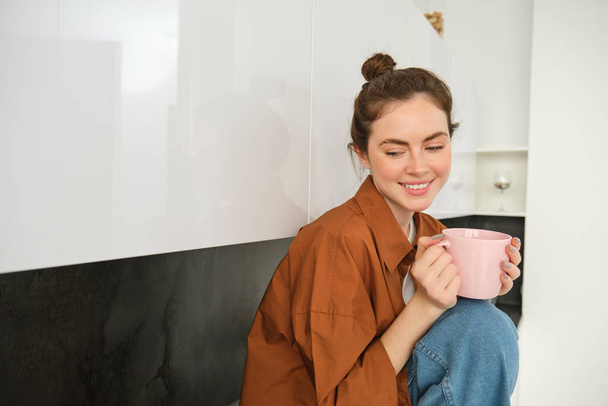 Bir fincan kahveyle genç bir kadının portresi, mutfakta oturuyor ve evde aromatik bir içecek içiyor, elinde çay fincanı tutuyor.. - Fotoğraf, Görsel