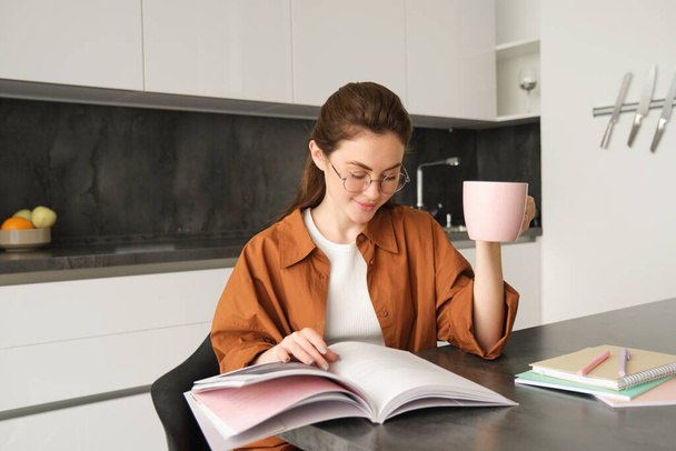 Portrait of beautiful student, woman in glasses reading and drinking coffee at home, studying and revising for lesson, doing homework. - 写真・画像