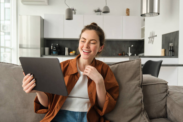 Portret kobiety śmiejącej się nad zabawnym filmem na tablecie cyfrowym, oglądającej film w serwisie streamingu gadżetów, siedzącej na kanapie w domu, czytającej e-booka. - Zdjęcie, obraz