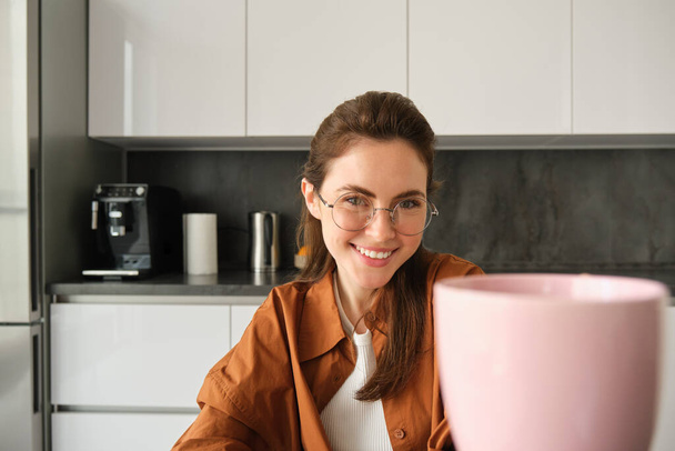 Mooie jonge vrouw in de keuken, die je een kopje thee geeft, thuis pauze neemt en koffie drinkt, een bril draagt, glimlacht naar de camera. - Foto, afbeelding