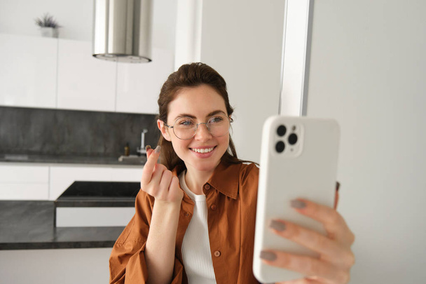 Nette junge Frau macht Selfie in ihrer Wohnung, zeigt Herzzeichen und macht mit Smartphone-Kamera schöne Bilder für die sozialen Medien. - Foto, Bild