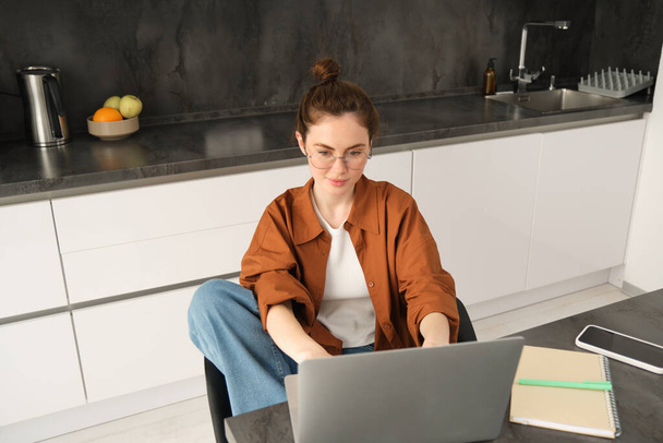 Portret pięknej młodej kobiety studiującej w domu, siedzącej z laptopem i dokumentami w kuchni. Samozatrudniony właściciel firmy wpisując na komputerze, wysyłając e-maile do klientów. - Zdjęcie, obraz