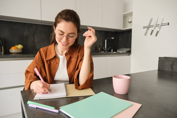 Portrait of young smiling woman, tutor preparing notes for lesson at home, writing in notebook and looking happy, student studying in kitchen. - Photo, Image
