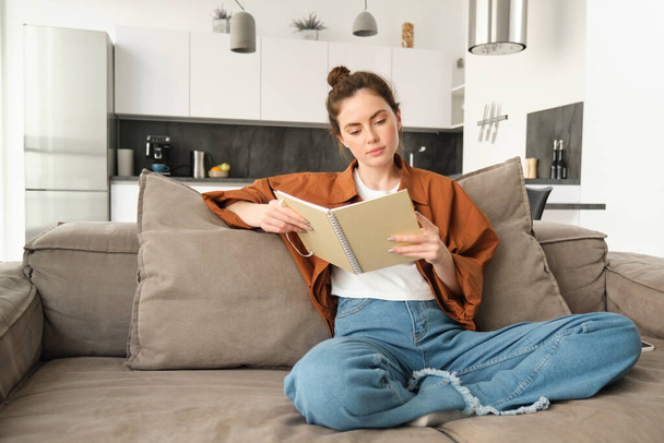 Bild einer Frau, die es sich auf dem Sofa bequem macht, sitzt und Notizbuch liest und ihre Notizen im Notizblock durchblättert. - Foto, Bild