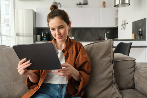 Porträt einer lächelnden schönen Frau, die auf dem Sofa sitzt, Videos auf einem digitalen Tablet anschaut, E-Books auf ihrem Gerät liest, sich zu Hause ausruht. - Foto, Bild