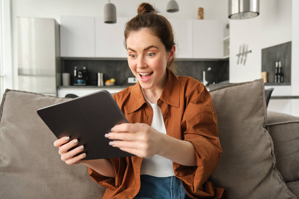 Portrét mladé ženy, která hraje videohry na digitálním tabletu, vypadá vzrušeně, naklápí gadget a dívá se na obrazovku, sedí na gauči v obývacím pokoji. - Fotografie, Obrázek