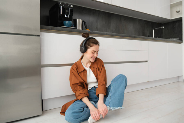 Linda chica moderna en el piso de la cocina en casa, escuchando música en auriculares inalámbricos, divirtiéndose. - Foto, imagen