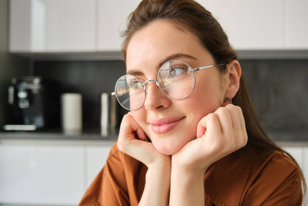 Portrait of lovely, happy young woman, wearing glasses, sitting in kitchen with dreamy, thoughtful face expression, spending time at home. - Photo, Image