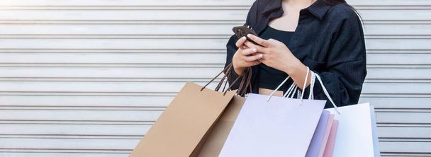 Mujer asiática usando smartphone y mirando hacia otro lado mientras disfruta de un día de compras. Viernes Negro venta y descuento. Comprar regalos de ropa para las vacaciones. - Foto, Imagen