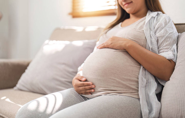 close-up, een mooie jonge vrouw tijdens het derde trimester van de zwangerschap Zwangere vrouw met armen op ronde buik, glimlachen, ontspannen uitdrukking.. - Foto, afbeelding