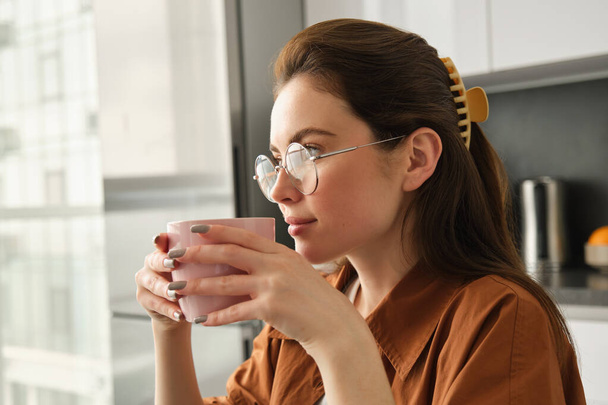 Portrait of young beautiful woman, relaxing with cup of fresh aroma coffee, holding mug and sitting in kitchen, smiling from happiness. - Photo, Image