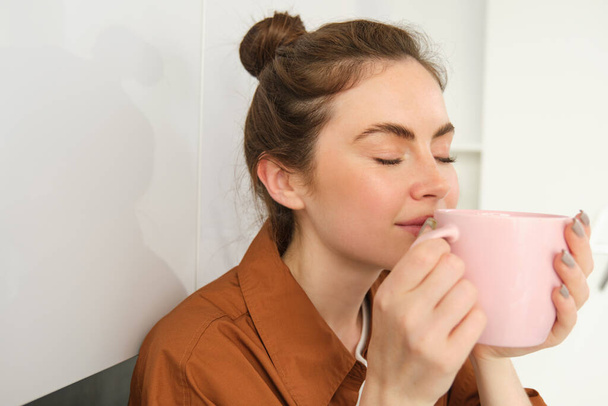 Zblízka portrét krásné brunetky žena s šálkem kávy, pití aromatický nápoj z hrnku, úsměv z potěšení, sedí v kuchyni doma. - Fotografie, Obrázek