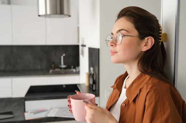 Portrait of cosy, beautiful and relaxed young woman at home, standing and leaning on wall with cup of coffee, drinking tea with thoughtful face expression. - Photo, Image