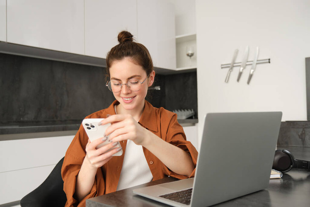 Porträt einer jungen Freiberuflerin, die von zu Hause aus mit Laptop arbeitet, Programmiererin, die mit Computer in der Küche sitzt, Anrufbeantworter auf dem Smartphone. - Foto, Bild