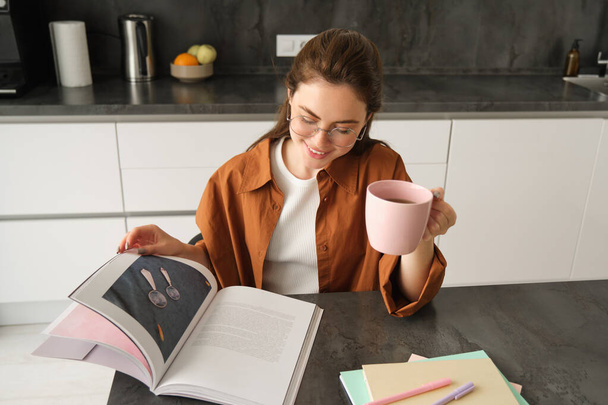 Retrato de una hermosa joven en la cocina, sentada con un libro, volteando páginas, leyendo y bebiendo café. - Foto, imagen