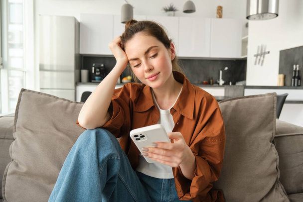 Retrato de una joven mujer moderna leyendo en el teléfono móvil, desplazándose aplicación de redes sociales en el teléfono inteligente, sentado en el sofá en la sala de estar con ropa casual. - Foto, imagen
