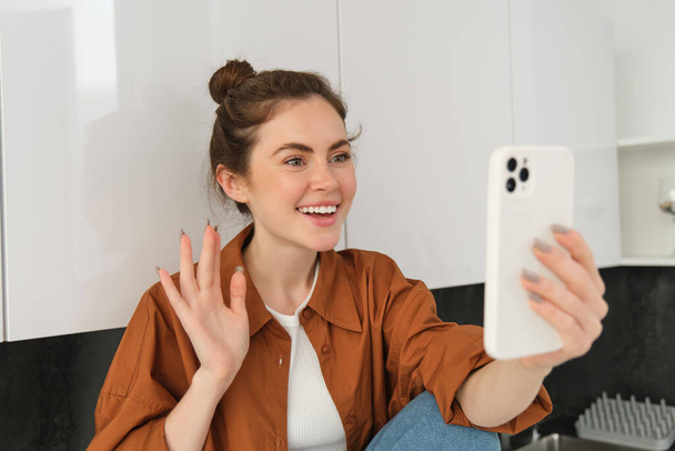 Portrait of young cheerful woman laughing and smiling during phone call, video chats with friend, sits on kitchen counter and talks to someone using smartphone app. - Photo, Image