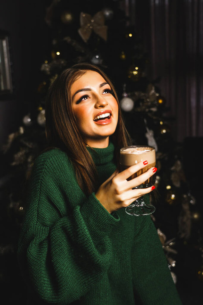 Kaunis nuori nainen vihreässä neulotussa puserossa, jossa on muki kuumaa kaakaota ja vaahtokarkkeja joulukuusen lähellä. Uusi vuosi - Valokuva, kuva