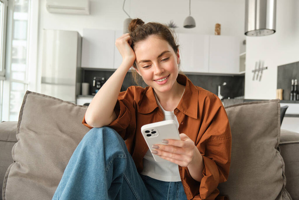 Portret van een gelukkige jonge brunette vrouw zittend op de bank met smartphone, het lezen van de melding, berichten op mobiele telefoon, rusten op de bank thuis, glimlachen terwijl het kijken naar het scherm van het apparaat. - Foto, afbeelding