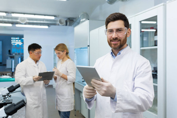 Portrait of mature experienced laboratory technician, laboratory worker smiling and looking at camera, man in glasses and protective gloves conducts experiments, uses tablet computer. - Photo, Image