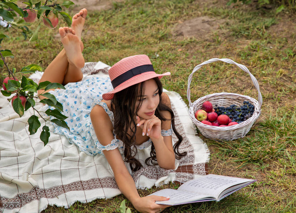 Beautiful young brunette caucasian woman wearing hat lying in the orchard garden and reading book. - Photo, Image