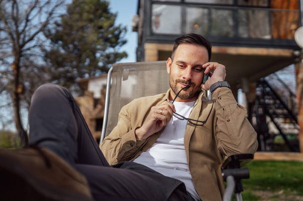 Jeden dorosły mężczyzna 40-letni biały mężczyzna siedzi przed swoim domem na wakacjach i rozmawia przez telefon komórkowy podczas rozmowy prawdziwa osoba kopiuje przestrzeń - Zdjęcie, obraz