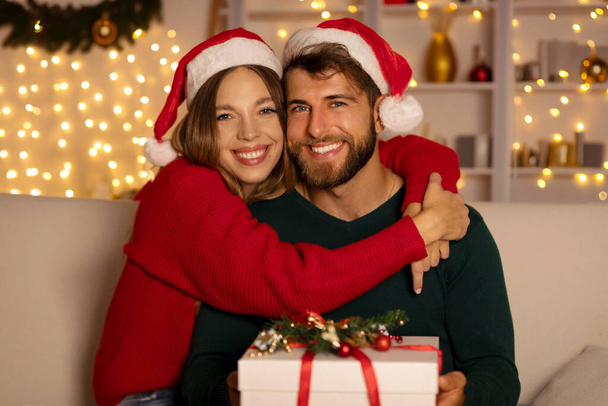 Szczęśliwa żona obejmująca męża, trzymając prezent na Boże Narodzenie, ciesząc się świętami Bożego Narodzenia, siedząc na kanapie w urządzonym pokoju z girlandami na tle - Zdjęcie, obraz