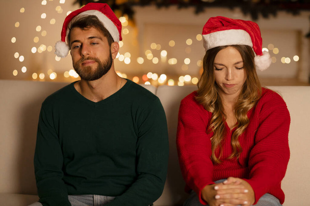 Relationships difficulties, winter holidays and people concept. Upset unhappy young couple sitting on sofa at home over garland lights on background - Photo, Image