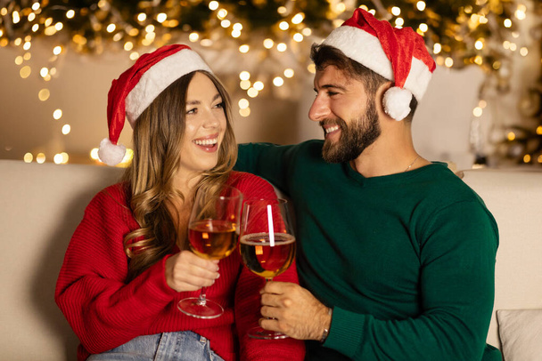 Любящие пары в шляпах Санта-Клауса веселятся с бокалами вина, наслаждаются отдыхом, сидят на диване в интерьере гостиной с елкой и светящимися гирляндами - Фото, изображение