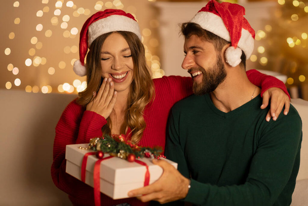 Romance and winter holidays. Surprised woman looks at gift box from husband, sitting in living room interior with Xmas tree with luminous garlands - Photo, Image