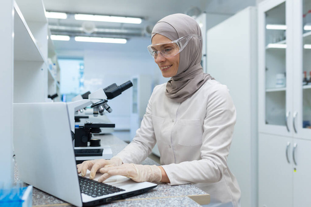 Arab fiatal nő tudós, gyógyszerész hidzsáb ül asztalnál mikroszkóppal, folyadékok üvegben és dolgozik laptop. - Fotó, kép