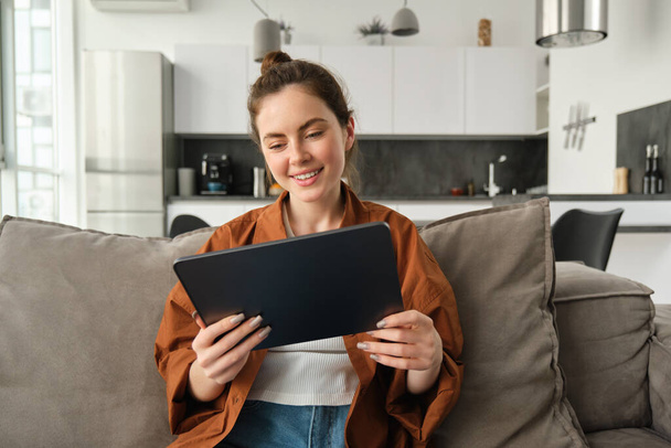 Portret van jonge brunette vrouw lezen op digitale tablet, tv-serie kijken op haar aanvraag, zitten op de bank in de woonkamer. - Foto, afbeelding