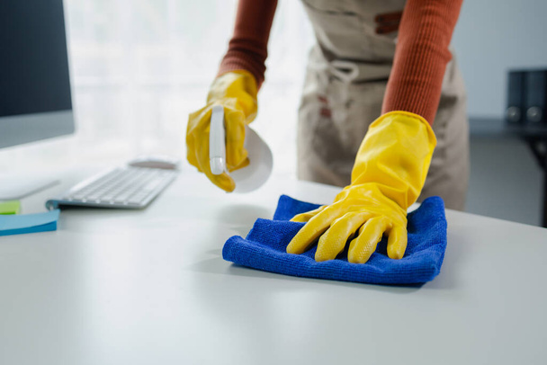 Καθαρισμός επιφάνεια γραφείο στο γραφείο με σπρέι απολυμαντικό, φορούν γάντια και σκουπίστε το τραπέζι με μια πετσέτα, η οικονόμος καθαρίζει το γραφείο εργασίας για την υγιεινή, λόγω της Covid-19, ιδέα καθαρισμού. - Φωτογραφία, εικόνα