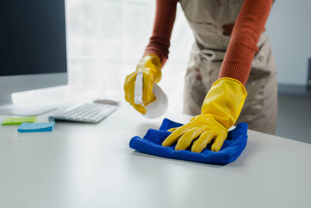 Ofisteki masa yüzeyini dezenfektan spreyiyle temizlemek, eldiven giymek ve masayı havluyla silmek, temizlikçi, temizlik için çalışma masasını temizliyor çünkü Covid-19, temizlik fikri.... - Fotoğraf, Görsel