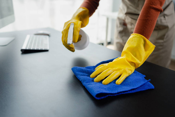 Sprzątanie powierzchni biurka w biurze sprayem do dezynfekcji, noszenie rękawiczek i wycieranie stołu ręcznikiem, gospodyni sprząta biurko dla higieny z powodu Covid-19, pomysł czyszczenia. - Zdjęcie, obraz