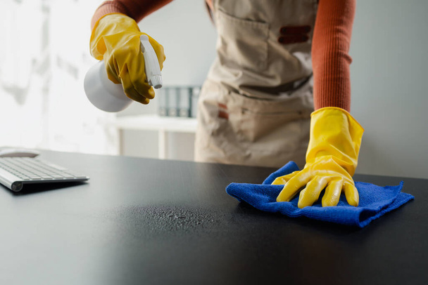 Cleaning desk surface in office with sanitizer spray, wear gloves and wipe the table with a towel, the housekeeper is cleaning the work desk for hygiene because of the Covid-19, cleaning idea. - Photo, Image
