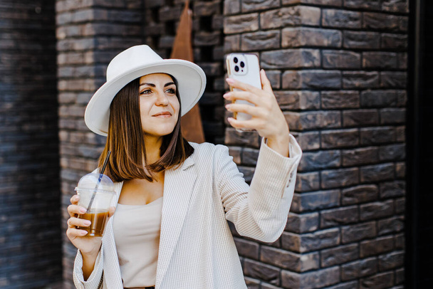 Beautiful woman going to meeting with cold coffee and holding the phone walking on the street. Portrait of successful business woman holding cup of cold drink in hand on her way to work on city street - Photo, Image
