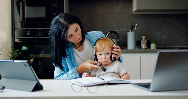 Woman holding baby infant and using computer laptop at home. Candid authentic and real life mom working and playing with son sitting on desk. - Φωτογραφία, εικόνα