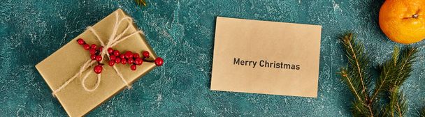 Wesołych Świąt koperta w pobliżu mandarynki i pudełko z jagodami ostrokrzewu na niebieskiej fakturze, baner - Zdjęcie, obraz