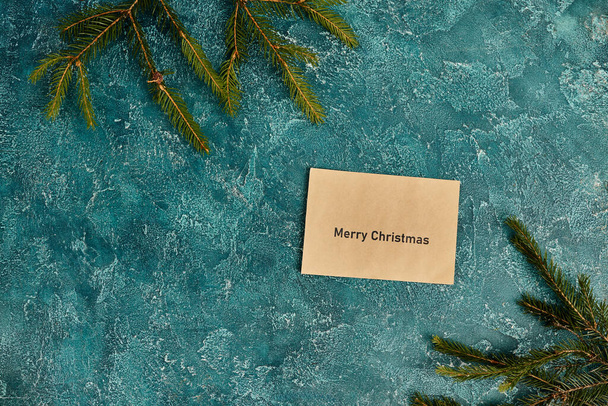 groet envelop met vrolijke kerst inscriptie in de buurt van dennenbomen takken op blauwe textuur achtergrond - Foto, afbeelding