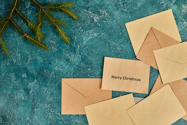 craft paper envelopes near green pine branches on blue textured backdrop, Merry Christmas lettering - Photo, Image